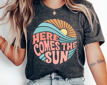 May include: Dark gray t-shirt with a circular graphic featuring a sun, waves, and the text "HERE COMES THE SUN" in pink and blue. The shirt is a casual, everyday wear item.