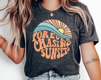 May include: Dark gray t-shirt with a circular graphic featuring a sunset design in orange, yellow, and blue. The text "Forever Chasing Sunsets" is arched around the design in orange. The shirt is paired with light blue jeans and white shoes.