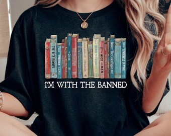 May include: Black t-shirt with the text "I'M WITH THE BANNED" and a graphic of a row of books. The person is sitting on a bed with a book, a bowl of food, and a black mug on a tray.