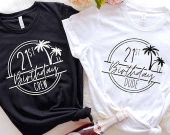 May include: Two t-shirts, one black and one white, with a round graphic that says "21st Birthday" and "Crew" or "Dude" with palm trees. The shirts are knotted at the bottom. A bottle of sparkling wine is in the foreground.