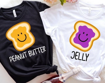 May include: Two t-shirts, one black and one white, each with a cartoon illustration of a slice of bread with a smiling face. The black shirt says "PEANUT BUTTER" and the white shirt says "JELLY".