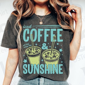 May include: Dark gray t-shirt with the words "iced COFFEE & SUNSHINE" in turquoise. The shirt features two iced coffee cup illustrations with straws. The phrase "all day every day" is also printed on the shirt.