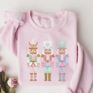 May include: A light pink long-sleeve sweatshirt featuring a graphic print of three nutcracker figures. The nutcrackers are decorated in pastel shades of mint green, light pink, and lavender with gold accents on their uniforms and hats.