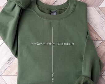 May include: A military green crewneck sweatshirt featuring a white vertical cross graphic. The text "THE WAY, THE TRUTH, AND THE LIFE" is printed horizontally across the center, with "JOHN 14:6" positioned vertically along the base of the cross.