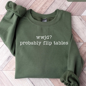 May include: Military green sweatshirt with the text "wwjd? probably flip tables" in white. The sweatshirt is made of a soft material and has a crew neck and long sleeves. The sleeves are cuffed at the wrists.