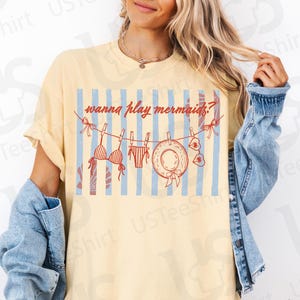 May include: A light yellow t-shirt with the text "wanna play mermaids?" above a graphic of a bikini, swimsuit, hat, and sunglasses hanging on a clothesline. The background features blue and white stripes. The person is wearing a denim jacket.