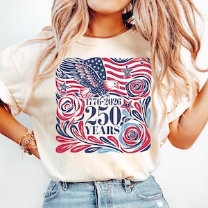 K&ouml;nnte beinhalten: Sandfarbenes T-Shirt mit patriotischem Design. Die Grafik zeigt eine amerikanische Flagge, einen Adler, Rosen und den Text "1776-2026 250 YEARS" in Rot, Wei&szlig; und Blau. Das Shirt ist ein l&auml;ssiger Stil.