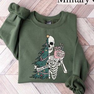 May include: A military green crewneck sweatshirt featuring a graphic print of a split figure that is half Christmas tree and half skeleton. The skeleton holds a pink mug with candy canes. A banner across the center reads "SORTA MERRY SORTA SCARY".