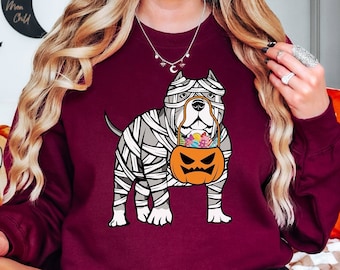 Könnte beinhalten: Ein bordeauxrotes Langarm-Sweatshirt mit einer Grafik eines Hundes, der wie eine Mumie in weiße Bandagen gewickelt ist und einen orangefarbenen Kürbis-Eimer mit bunten Süßigkeiten im Maul hält, vor einem dunkelroten Hintergrund.