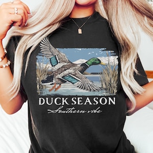 May include: Black t-shirt featuring a graphic of a flying duck over a lake with reeds. The text "DUCK SEASON Southern vibe" is printed below the image. The shirt is a comfortable, casual style.