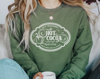 May include: An olive green crewneck sweatshirt featuring a white graphic print that reads Old Fashioned Hot Cocoa Served Here North Pole Warm Up Here Open All Winter, accompanied by an illustration of a mug with a candy cane.