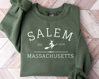 May include: An olive green crewneck sweatshirt featuring white screen-printed text that reads SALEM, EST. 1626, and MASSACHUSETTS. A graphic of a witch flying on a broomstick is positioned between the established date and a horizontal line.