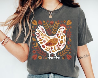 May include: A charcoal gray short-sleeve t-shirt featuring a central graphic of a white chicken filled with a pattern of autumn-colored leaves, acorns, and flowers in shades of orange, red, brown, and yellow, set against a dark fabric background.