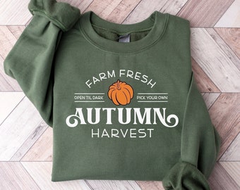 May include: An olive green crewneck sweatshirt featuring a white graphic print that reads FARM FRESH, OPEN TIL DARK, PICK YOUR OWN, AUTUMN, and HARVEST, centered around an orange pumpkin illustration with a dark green stem and defined ridges.