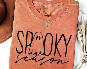 May include: A burnt orange short-sleeve t-shirt featuring a black graphic of a ghost and the text "SPOOKY season" in a mix of block and script lettering. The shirt has a crew neckline and a Comfort Colors brand label visible at the inner collar.