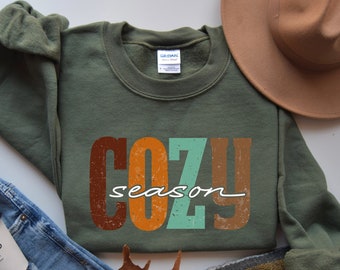 May include: A military green crewneck sweatshirt featuring the text COZY in large, distressed, multi-colored block letters and the word season in a white cursive script font. The garment has a Gildan brand label at the inner neckline.