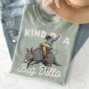 May include: A light green t-shirt with the text "KIND OF A Big Dillo" and a graphic of a cowboy riding an armadillo. The shirt is laid on top of a pair of light blue jeans, a tan hat, and a gold necklace.