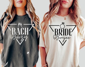 May include: Two oversized t-shirts, one charcoal gray and one cream, with black text and geometric designs. The gray shirt says "Bach & Boozy" with champagne glasses, and the cream shirt says "Bride & Boujee" with a diamond ring.