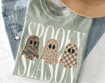 Könnte beinhalten: Ein salbeigrünes T-Shirt mit dem Schriftzug SPOOKY SEASON in weißen Buchstaben und drei Geistergrafiken mit Camouflage-, Leoparden- und Schachbrettmuster. Das Shirt liegt auf einer hellblauen Jeans, einem beigen Fedora-Hut und einer silbernen Kette.