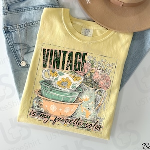 May include: A yellow t-shirt with the text "VINTAGE is my favorite color" and a graphic of teacups and flowers. The shirt is laid on top of blue jeans, with a necklace and a tan hat in the background.
