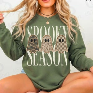 May include: A military green long-sleeve sweatshirt featuring the white text SPOOKY SEASON and three ghost illustrations. The ghosts have different patterns: one with camouflage, one with leopard print, and one with a brown and white checkerboard pattern.