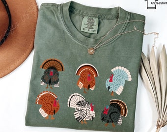 May include: An olive green crew neck t-shirt featuring a graphic print of six turkeys in various colors including brown, white, and light blue. The shirt has a Comfort Colors brand label on the inside collar and a small logo in the top right corner.