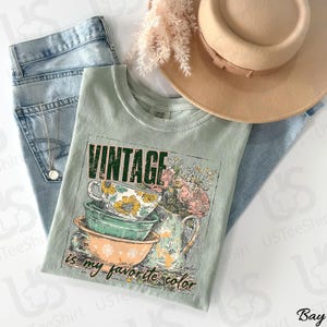 May include: A sage green t-shirt with the text "VINTAGE is my favorite color" and a graphic of teacups and flowers. The shirt is paired with light wash jeans, a gold necklace, and a tan hat.
