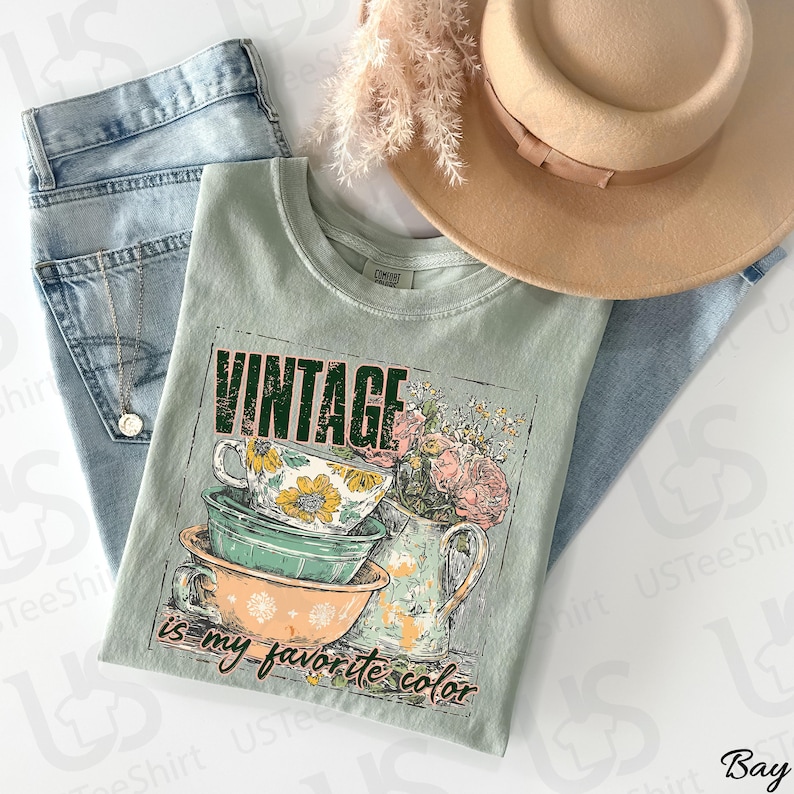 May include: A sage green t-shirt with the text "VINTAGE is my favorite color" and a graphic of teacups and flowers. The shirt is paired with light wash jeans, a gold necklace, and a tan hat.