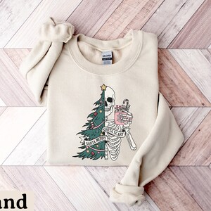 May include: A sand-colored crewneck sweatshirt featuring a graphic design split down the middle. One side shows a green Christmas tree with ornaments, while the other side shows a skeleton holding a pink mug with a candy cane. Text reads Sorta Merry Sorta Scary.