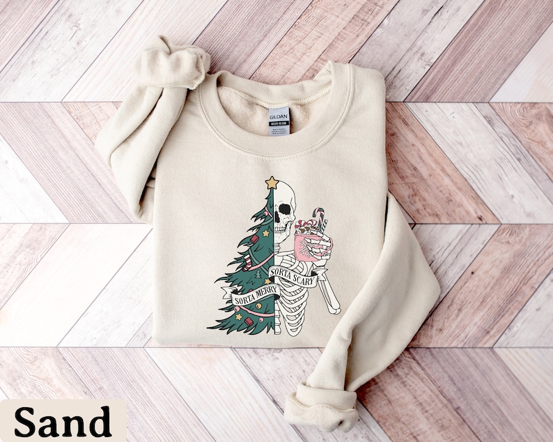 May include: A sand-colored crewneck sweatshirt featuring a graphic design split down the middle. One side shows a green Christmas tree with ornaments, while the other side shows a skeleton holding a pink mug with a candy cane. Text reads Sorta Merry Sorta Scary.