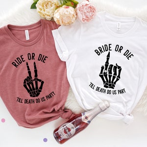 Puede incluir: Dos camisetas, una malva y otra blanca, con texto negro y gr&aacute;ficos de manos de esqueleto. La camiseta malva dice "RIDE OR DIE" con un gesto de la mano de la paz. La camiseta blanca dice "BRIDE OR DIE" con un gesto de dedo medio.
