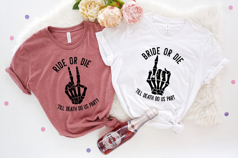 Puede incluir: Dos camisetas, una malva y otra blanca, con texto negro y gr&aacute;ficos de manos de esqueleto. La camiseta malva dice "RIDE OR DIE" con un gesto de la mano de la paz. La camiseta blanca dice "BRIDE OR DIE" con un gesto de dedo medio.