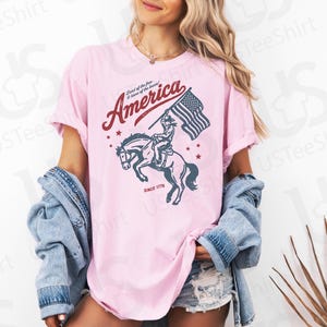 Puede incluir: Camiseta rosa claro con un gr&aacute;fico de un jinete sobre un caballo encabritado sosteniendo una bandera estadounidense. La palabra "America" est&aacute; arqueada sobre la imagen en color burdeos, con el texto "Land of the free & home of the brave" encima. El texto "Since 1776" est&aacute; debajo.