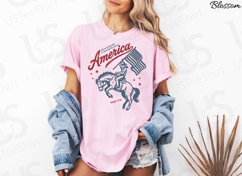 Puede incluir: Camiseta rosa claro con un gr&aacute;fico de un jinete sobre un caballo encabritado sosteniendo una bandera estadounidense. La palabra "America" est&aacute; arqueada sobre la imagen en color burdeos, con el texto "Land of the free & home of the brave" encima. El texto "Since 1776" est&aacute; debajo.