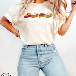 Puede incluir: Una camiseta de manga corta color crema con un estampado gr&aacute;fico de cuatro porciones de tarta distintas dispuestas en una fila horizontal. La ilustraci&oacute;n incluye una tarta de cerezas roja, una tarta de calabaza naranja con nata, una tarta de nueces marr&oacute;n y una tarta de manzana amarilla.