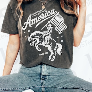 Puede incluir: Camiseta gris oscuro con un gr&aacute;fico blanco de un jinete a caballo sosteniendo una bandera estadounidense. El texto "America" est&aacute; encima del gr&aacute;fico, con "Land of the free & home of the brave!" y "SINCE 1776" tambi&eacute;n impresos.
