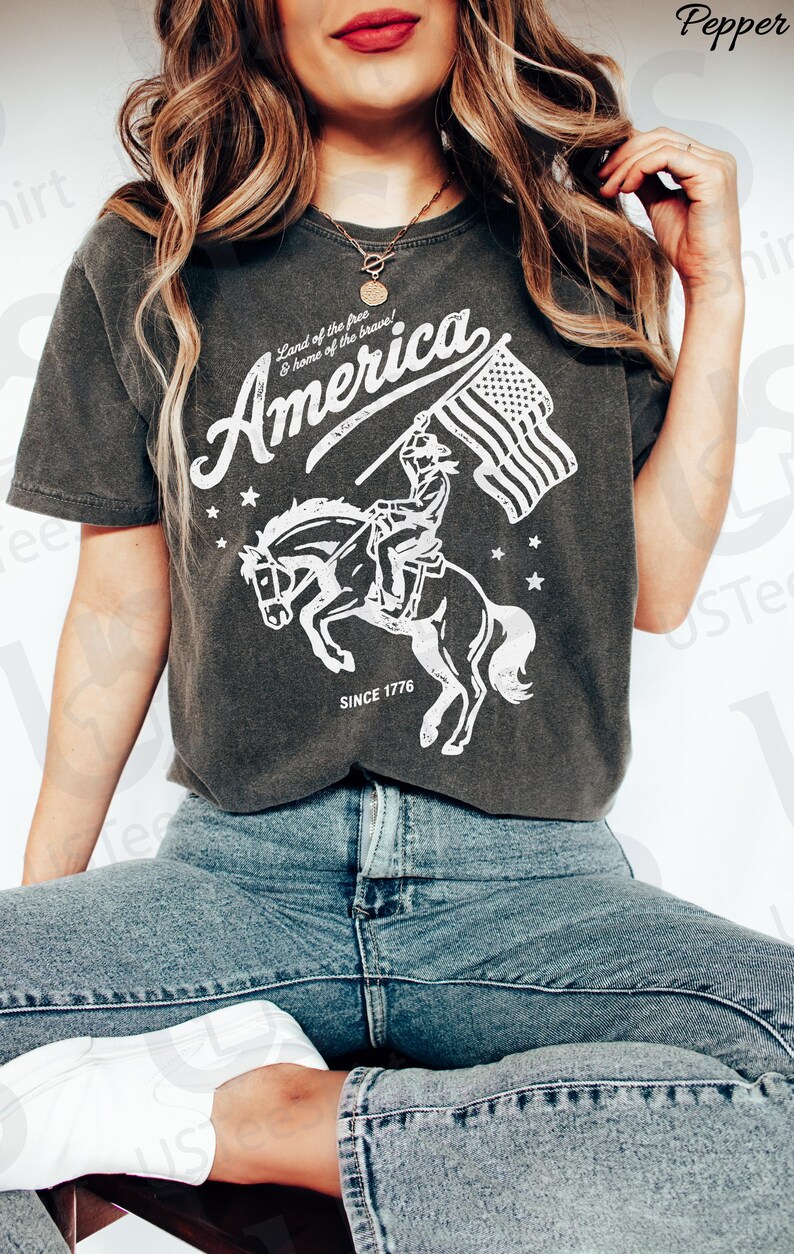 Puede incluir: Camiseta gris oscuro con un gr&aacute;fico blanco de un jinete a caballo sosteniendo una bandera estadounidense. El texto "America" est&aacute; encima del gr&aacute;fico, con "Land of the free & home of the brave!" y "SINCE 1776" tambi&eacute;n impresos.