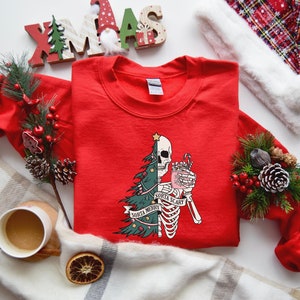 May include: A red crewneck sweatshirt featuring a graphic print of a split figure that is half a decorated Christmas tree and half a skeleton holding a pink mug with a candy cane. The text on the graphic reads Sorta Merry and Sorta Scary.