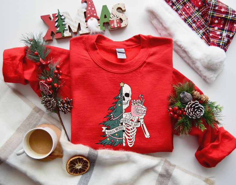 May include: A red crewneck sweatshirt featuring a graphic print of a split figure that is half a decorated Christmas tree and half a skeleton holding a pink mug with a candy cane. The text on the graphic reads Sorta Merry and Sorta Scary.