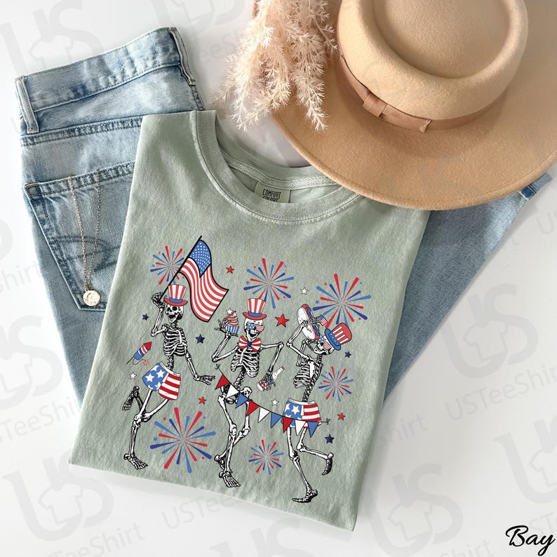 May include: A light green t-shirt featuring a festive design with three skeleton figures celebrating with fireworks, flags, and patriotic hats. The shirt is paired with blue jeans and a tan hat, perfect for a patriotic holiday.