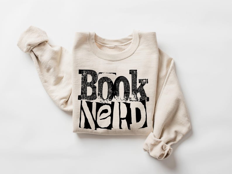 May include: A cream-coloured sweatshirt featuring the text "Book Nerd" in large, distressed black lettering. The garment is folded with rolled-up sleeves, set against a white backdrop.