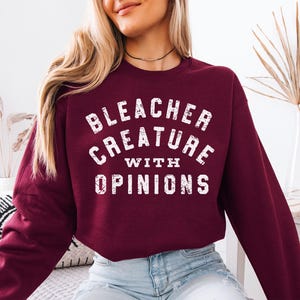 Puede incluir: Sudadera burdeos con la frase blanca "BLEACHER CREATURE WITH OPINIONS". La tipograf&iacute;a es desgastada. La modelo lleva vaqueros azules. La sudadera es de color liso.