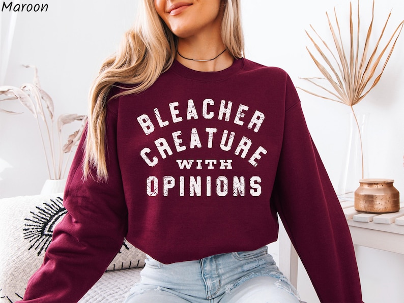 Puede incluir: Sudadera burdeos con la frase blanca "BLEACHER CREATURE WITH OPINIONS". La tipograf&iacute;a es desgastada. La modelo lleva vaqueros azules. La sudadera es de color liso.