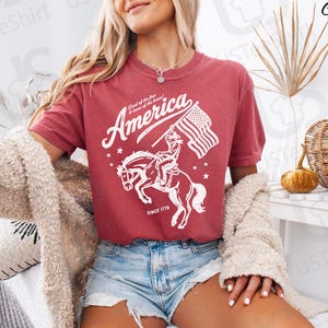 Puede incluir: Camiseta de color carmes&iacute; con un gr&aacute;fico blanco de un jinete a caballo sosteniendo una bandera estadounidense. La palabra "America" est&aacute; arqueada sobre la imagen, con "Land of the free & home of the brave" y "Since 1776" debajo.