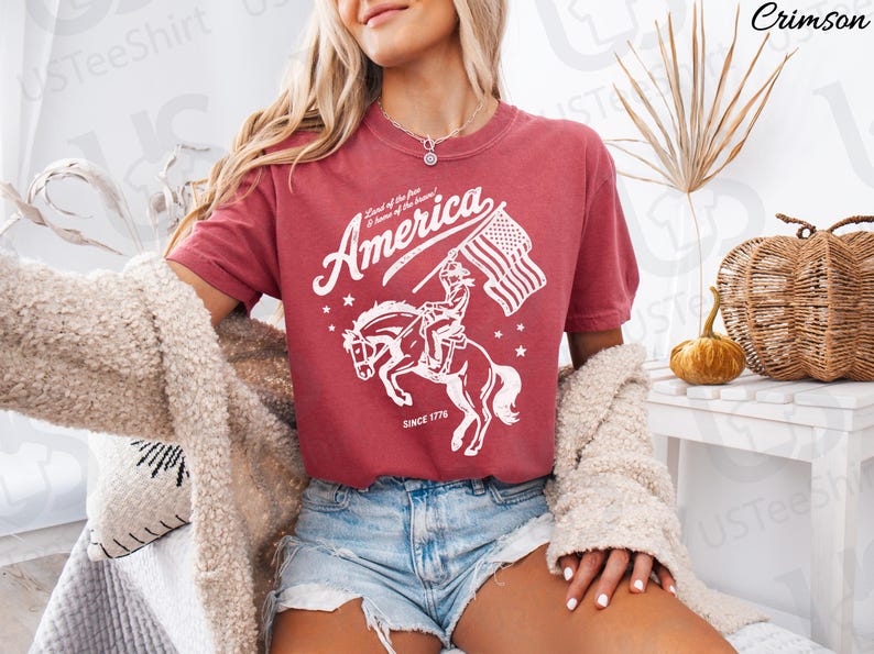 Puede incluir: Camiseta de color carmes&iacute; con un gr&aacute;fico blanco de un jinete a caballo sosteniendo una bandera estadounidense. La palabra "America" est&aacute; arqueada sobre la imagen, con "Land of the free & home of the brave" y "Since 1776" debajo.