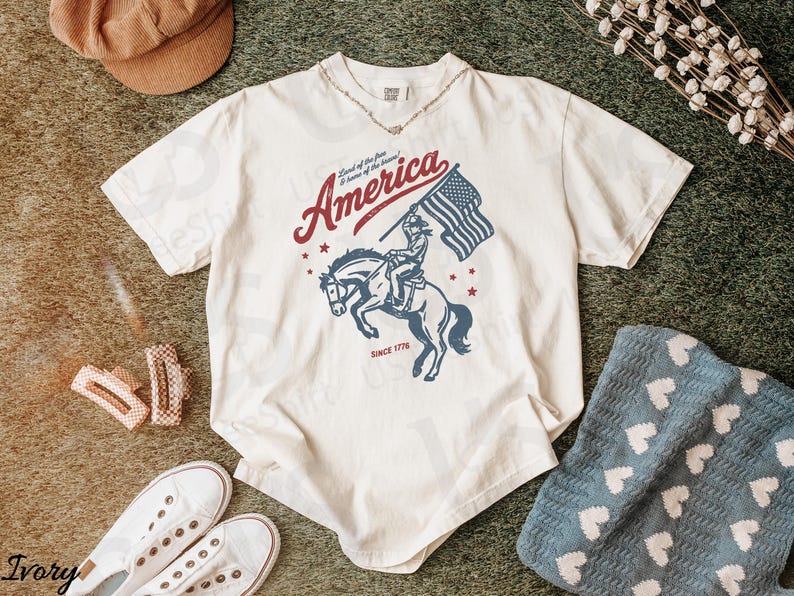 Puede incluir: Camiseta de color crema con un gr&aacute;fico desgastado de un jinete a caballo sosteniendo una bandera estadounidense, con la palabra "America" y el texto "Since 1776". La camiseta est&aacute; rodeada de accesorios.
