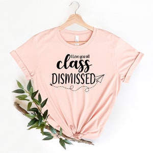 May include: A light pink t-shirt with the text "I love you all class DISMISSED" in black script. A paper airplane graphic and dotted line trail are below the text. The shirt is hanging on a wooden hanger.