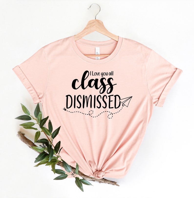 May include: A light pink t-shirt with the text "I love you all class DISMISSED" in black script. A paper airplane graphic and dotted line trail are below the text. The shirt is hanging on a wooden hanger.