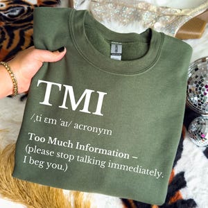 May include: Olive green sweatshirt with the white text "TMI" and its definition: "Too Much Information - (please stop talking immediately. I beg you.)". The sweatshirt has a crew neck and long sleeves.