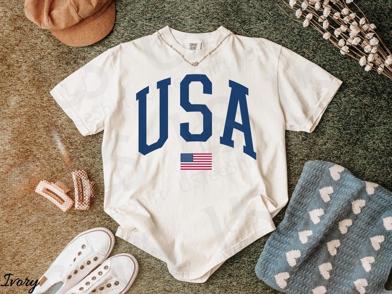 May include: Cream-colored t-shirt featuring the word "USA" in navy blue block letters and a small American flag. The shirt is styled with white trainers, a brown cap, and hair clips.